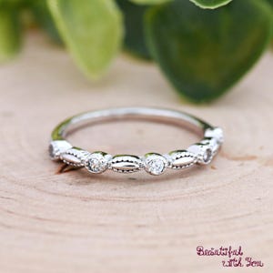 May include: A silver ring with small, clear gemstones set in a repeating pattern. The ring is on a wooden surface, with green foliage in the background. The ring has a delicate, elegant design.