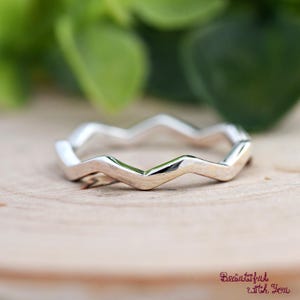 May include: A silver-colored ring with a zigzag design. The ring is set against a wooden surface with green foliage in the background. The ring's design is a series of connected chevrons.
