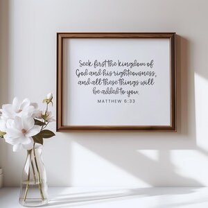 This mockup shows what this unframed Matthew 6 33 Bible verse poster could look like in a wood frame. It has a white background and gray text. It says seek first the kingdom of God ... It is shown in a minimalist and modern room.
