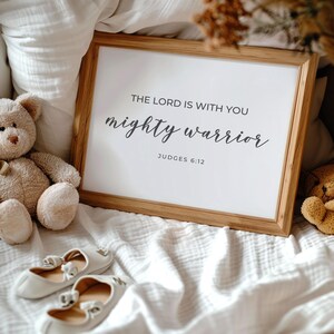 May include: A framed print with a white background and black text that reads "The Lord is with you mighty warrior Judges 6:12". The print is sitting on a white bedspread with a teddy bear and a pair of white shoes.