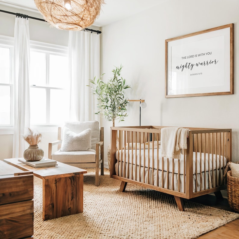 May include: A light wood cot with a white mattress and a white blanket. The cot is in a nursery with a light wood coffee table, a white armchair, and a potted plant. A framed print with the text "The Lord is with you mighty warrior" hangs on the wall.