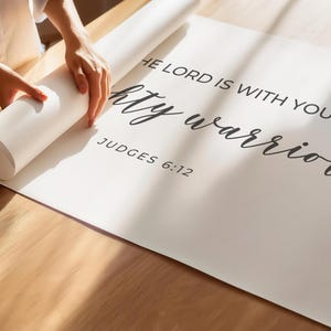 May include: White poster with black text that reads "The Lord is with you mighty warrior" and "Judges 6:12". A person is rolling up the poster.