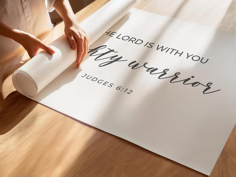 May include: White poster with black text that reads "The Lord is with you mighty warrior" and "Judges 6:12". A person is rolling up the poster.
