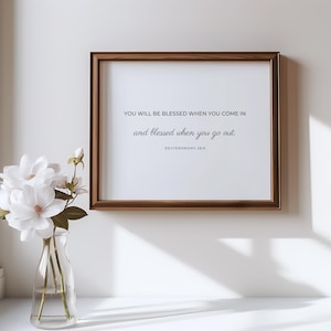 May include: A framed print with a white background and a brown wooden frame. The print features a quote from Deuteronomy 28:6, "You will be blessed when you come in and blessed when you go out." The print is displayed on a white wall with a vase of white flowers in front of it.