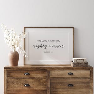 May include: A framed print with a white background and black text that reads "The Lord is with you mighty warrior Judges 6:12". The print is on a wooden dresser with a vase of white flowers.