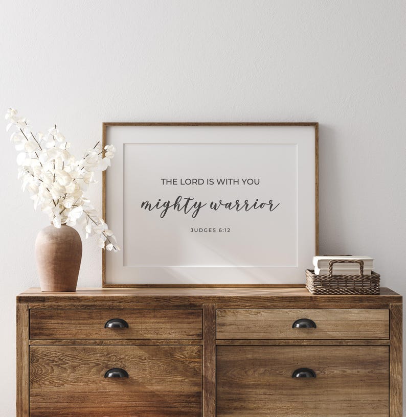 May include: A framed print with a white background and black text that reads "The Lord is with you mighty warrior Judges 6:12". The print is on a wooden dresser with a vase of white flowers.