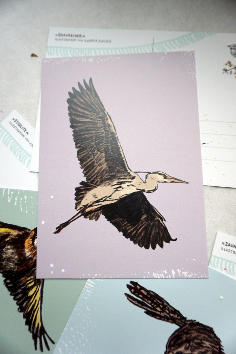 May include: A close-up of a postcard featuring a detailed illustration of a grey heron in flight against a light purple background. The heron has brown and black markings on its wings and body. The postcard is decorated with white dots.