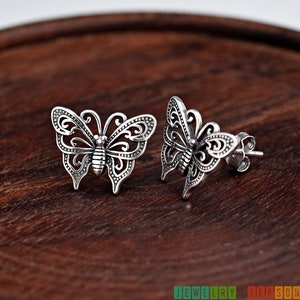 May include: A pair of silver butterfly stud earrings. The earrings feature intricate openwork designs on the wings and body. The earrings are displayed on a dark wooden surface. The text "JEWELRY SEASON" is visible in the bottom right corner.