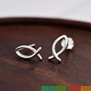 May include: A pair of silver-colored stud earrings in the shape of the Christian fish symbol. The earrings are displayed on a dark brown wooden surface. The earrings are small and minimalist.