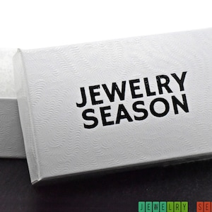 K&ouml;nnte beinhalten: Wei&szlig;e Schmuckschatulle mit einem strukturierten Deckel und dem Aufdruck "Jewelry Season" in Schwarz.