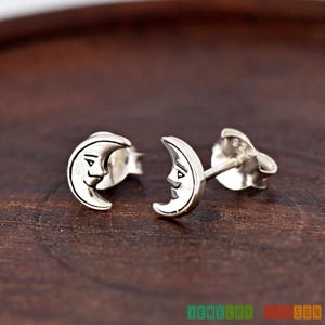 May include: A pair of silver crescent moon earrings with a face on each moon. The earrings are on a wooden surface.
