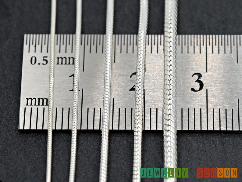 K&ouml;nnte beinhalten: Vier silberne Schlangenketten in verschiedenen Breiten liegen neben einem Lineal. Die Ketten werden in Millimetern gemessen, wobei die kleinste Kette 1 mm und die gr&ouml;&szlig;te Kette 3 mm misst.