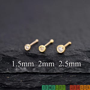 May include: Three gold nose studs with clear, round gemstones. The studs are displayed on a dark, textured surface. The text below the studs reads "1.5mm 2mm 2.5mm". The bottom right corner of the image has the text "JEWELRY SEASON".