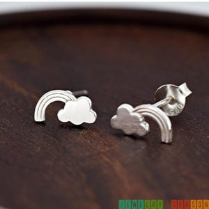 May include: A pair of silver stud earrings featuring a rainbow and cloud design. The earrings are made of a shiny, metallic material and are set against a dark brown background. The earrings are small and delicate, perfect for everyday wear.