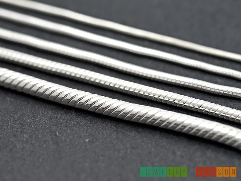 K&ouml;nnte beinhalten: Nahaufnahme einer silbernen Schlangenkettenhalskette. Die Kette besteht aus kleinen, ineinandergreifenden Gliedern, die einen glatten, flie&szlig;enden Look erzeugen.