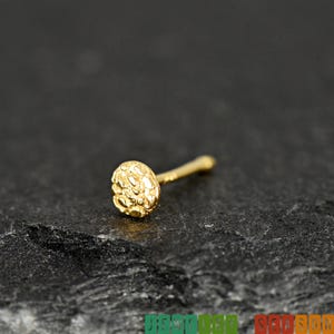 May include: A gold nose stud featuring a circular head with a textured, uneven surface. The stud has a thin, straight post extending from the back of the circular head. The piece is rendered in a polished gold tone with a distinct, rugged finish.