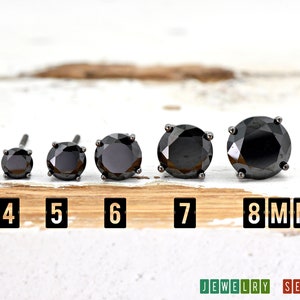 May include: Five black round stud earrings in different sizes, ranging from 4mm to 8mm. The earrings are displayed on a wooden surface with the size of each earring labeled below it.