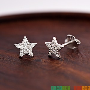 May include: A pair of star-shaped stud earrings. Each earring features a five-pointed star design, encrusted with small, round, clear gemstones. The earrings are set on silver posts. The background is a dark, wooden surface.