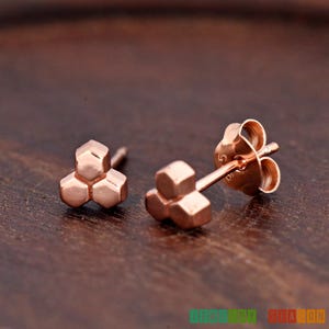 May include: A pair of rose gold stud earrings with a geometric design. Each earring features three connected hexagonal shapes.