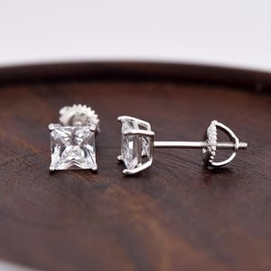 May include: A pair of square-cut, clear cubic zirconia stud earrings. The earrings are set in a silver-colored metal with a post and screw-back closure. The earrings are displayed on a dark brown wooden surface.