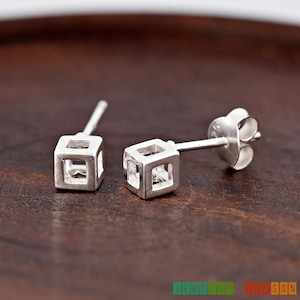May include: A pair of silver cube stud earrings with a hollow center. The earrings are on a wooden surface.