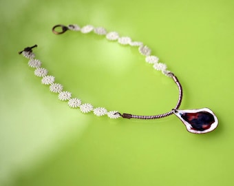 May include: A white crocheted necklace with a small, teardrop-shaped pendant. The pendant is made of metal and has a red and black design.