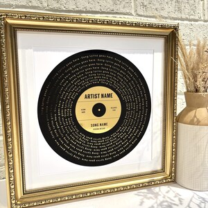 May include: A framed black and gold record with the song title "SONG NAME" and the artist name "ARTIST NAME" printed in gold lettering. The record is surrounded by a gold frame.