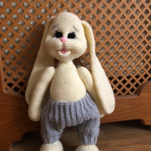 May include: A crocheted bunny toy with long ears, wearing gray pants. The bunny has a pink nose, black eyes, and a white and gray face.
