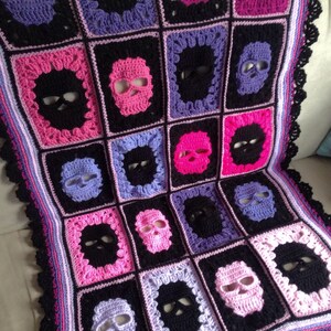 May include: Crocheted blanket featuring a repeating pattern of skull motifs. The blanket is made with black, pink, purple, and lavender yarn. The edges are finished with a black, scalloped border. The blanket is a square shape.