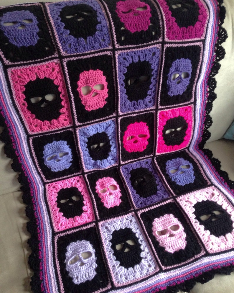 May include: Crocheted blanket featuring a repeating pattern of skull motifs. The blanket is made with black, pink, purple, and lavender yarn. The edges are finished with a black, scalloped border. The blanket is a square shape.