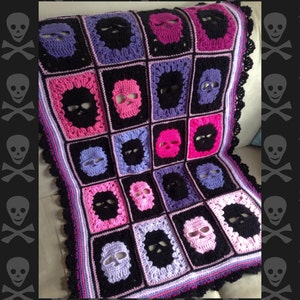 May include: Crocheted blanket featuring a repeating skull pattern in black, pink, purple, and white. The blanket has a black border with a scalloped edge. The skulls are centered in black squares, creating a unique and eye-catching design.