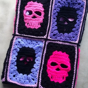 May include: A crocheted blanket featuring four squares, each with a skull design. The skulls are in pink and black, set against a black background, with purple and pink borders. The blanket is handmade.