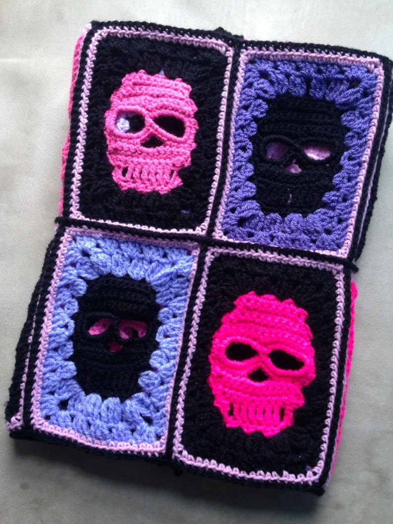 May include: A crocheted blanket featuring four squares, each with a skull design. The skulls are in pink and black, set against a black background, with purple and pink borders. The blanket is handmade.
