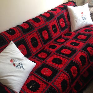 May include: A crocheted blanket in a square pattern, featuring red and black skull motifs. The blanket is draped over a sofa, accompanied by two white throw pillows with skull and rose embroidery. The color scheme is primarily red, black, and gray.