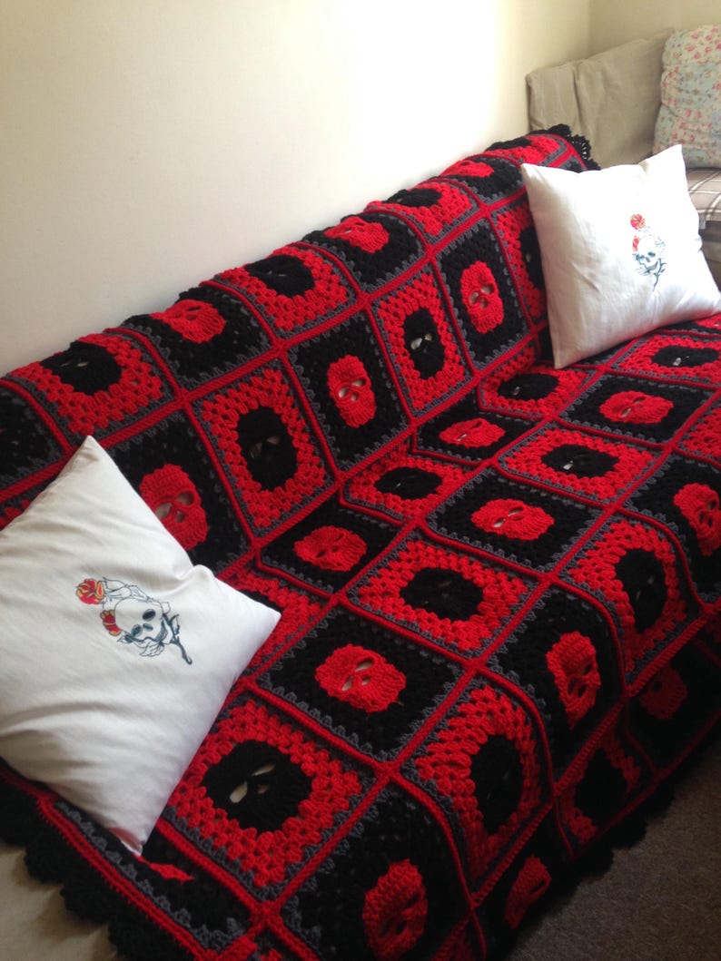 May include: A crocheted blanket in a square pattern, featuring red and black skull motifs. The blanket is draped over a sofa, accompanied by two white throw pillows with skull and rose embroidery. The color scheme is primarily red, black, and gray.