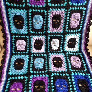 May include: Crocheted blanket featuring a repeating skull pattern in black, purple, blue, and light blue. The blanket has a black border with a scalloped edge and a striped border in shades of purple and blue.