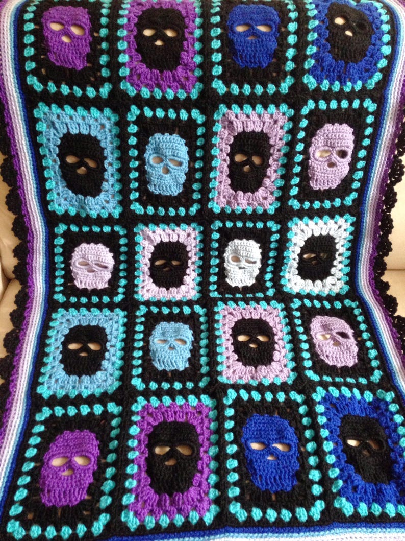 May include: Crocheted blanket featuring a repeating skull pattern in black, purple, blue, and light blue. The blanket has a black border with a scalloped edge and a striped border in shades of purple and blue.