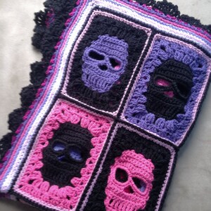May include: A crocheted blanket featuring four square panels with skull designs. The skulls are in purple, black, and pink, set against a black background. The blanket has a black scalloped edge and a border of purple, pink, and white.