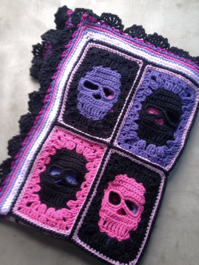 May include: A crocheted blanket featuring four square panels with skull designs. The skulls are in purple, black, and pink, set against a black background. The blanket has a black scalloped edge and a border of purple, pink, and white.