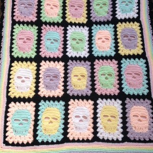 May include: A handmade crochet blanket featuring a repeating pattern of colorful skulls on a black background. The skulls are in pastel shades of pink, blue, yellow, and green, framed by a white border. The blanket has a rainbow border and a black scalloped edge.