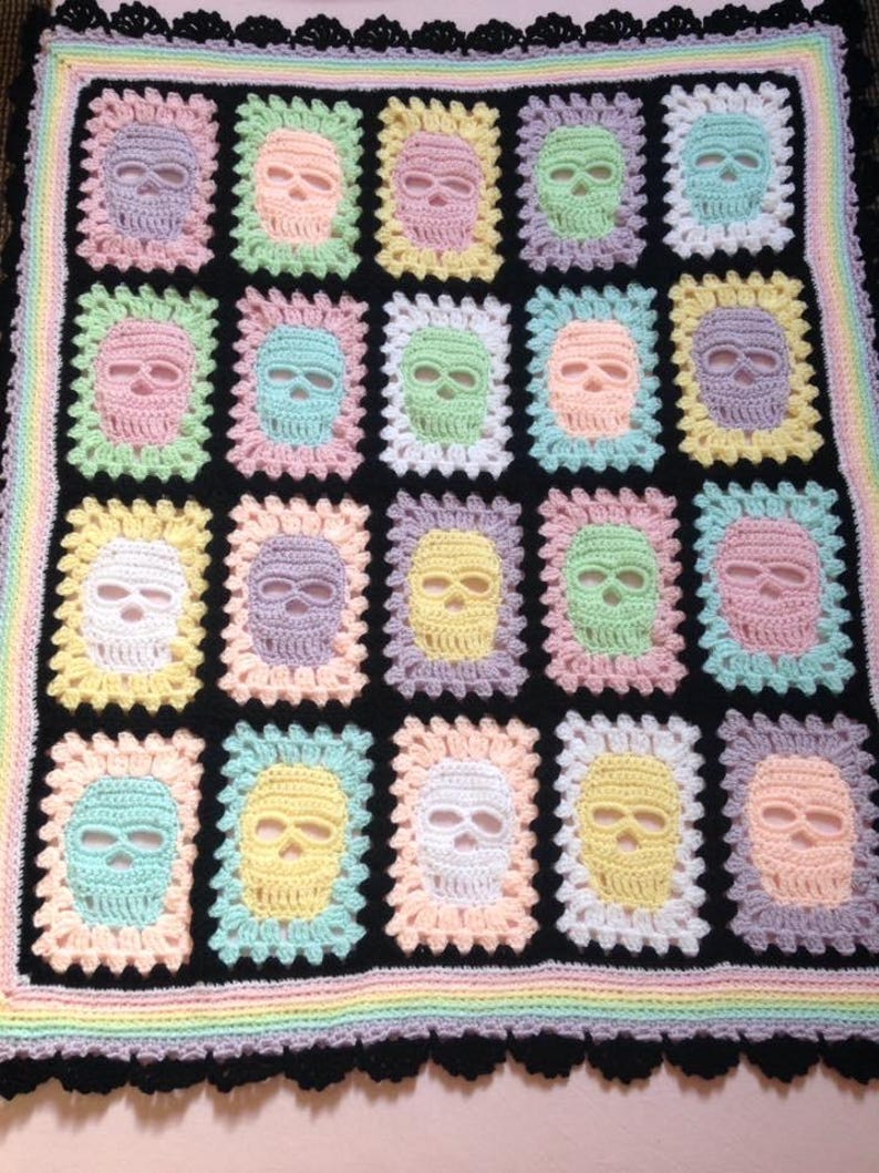May include: A handmade crochet blanket featuring a repeating pattern of colorful skulls on a black background. The skulls are in pastel shades of pink, blue, yellow, and green, framed by a white border. The blanket has a rainbow border and a black scalloped edge.