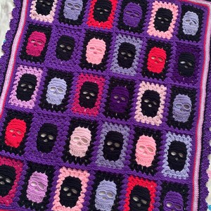 May include: A crocheted blanket featuring a repeating pattern of skull motifs in various colors. The skulls are framed in contrasting colors against a purple background. The blanket has a scalloped edge.