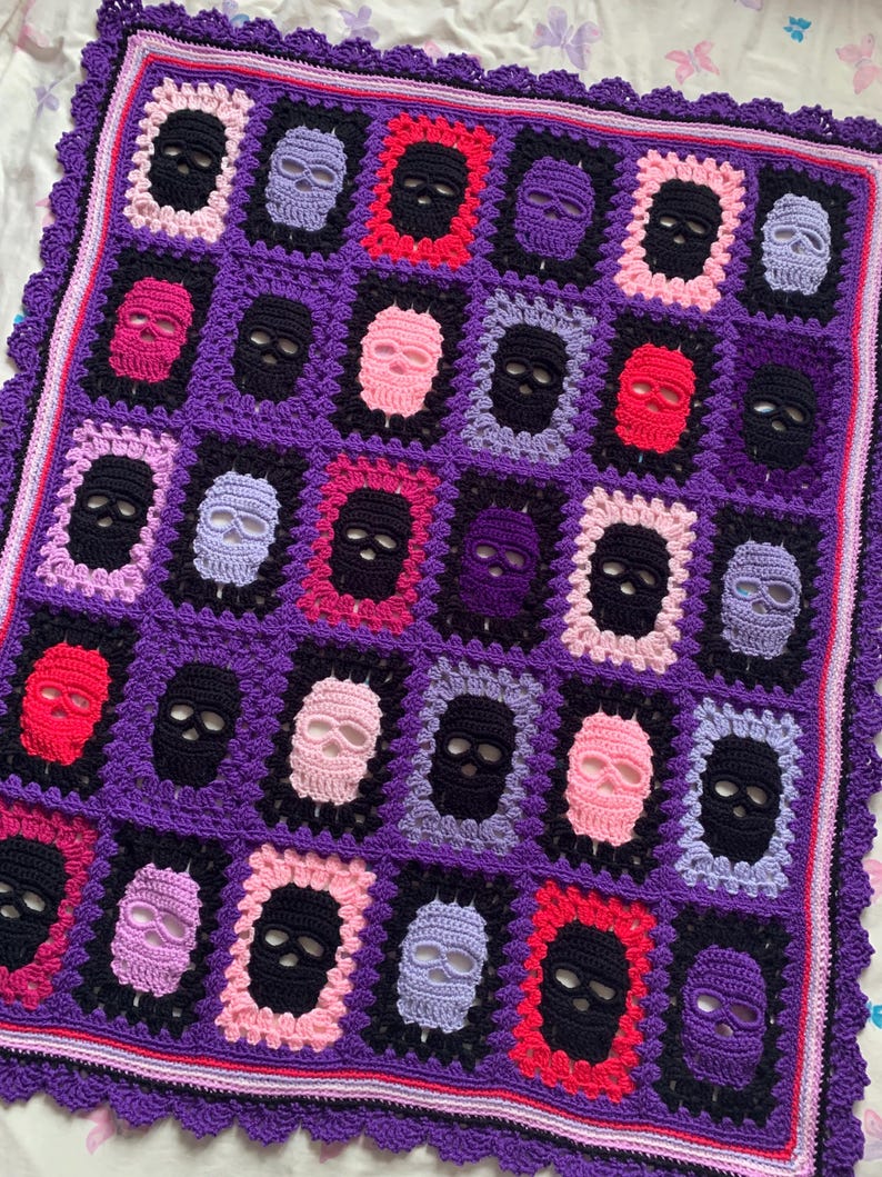 May include: A crocheted blanket featuring a repeating pattern of skull motifs in various colors. The skulls are framed in contrasting colors against a purple background. The blanket has a scalloped edge.