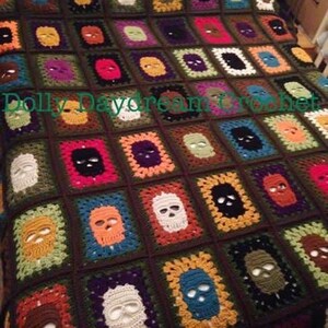 May include: A handmade crochet blanket featuring a repeating skull pattern. Each square showcases a skull in various colors, including white, orange, and blue, set against a dark brown background. The blanket is a unique, cozy home decor item.
