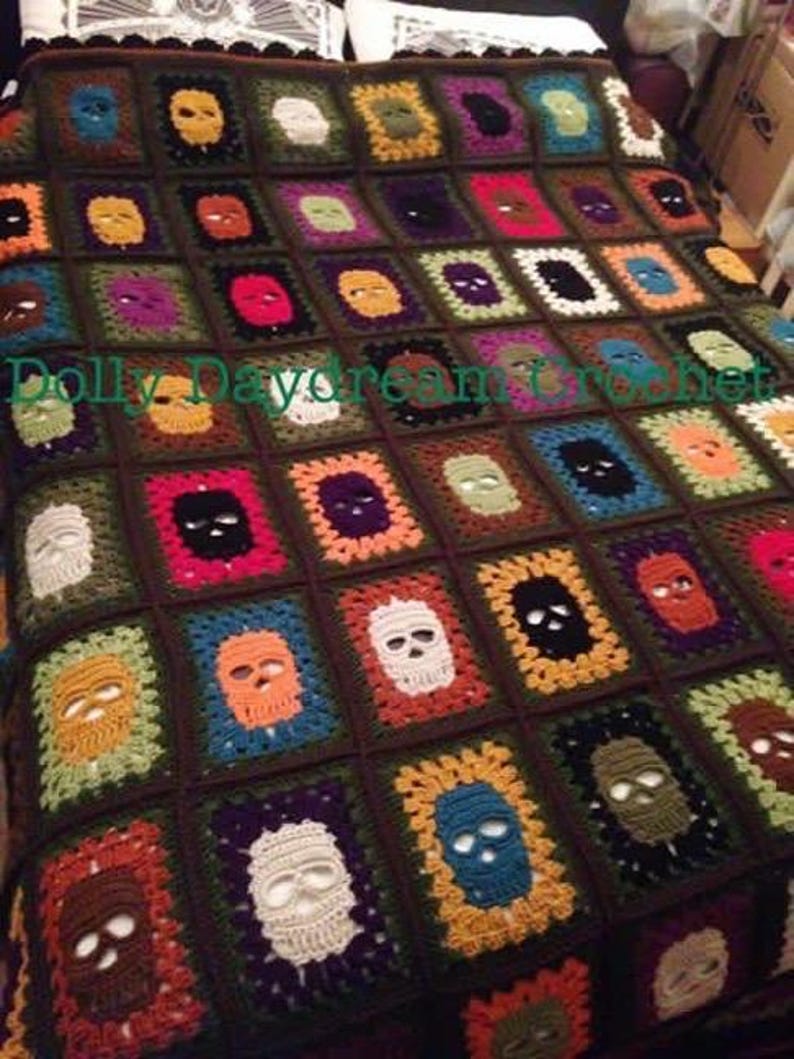 May include: A handmade crochet blanket featuring a repeating skull pattern. Each square showcases a skull in various colors, including white, orange, and blue, set against a dark brown background. The blanket is a unique, cozy home decor item.