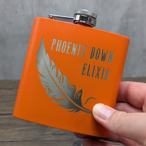 May include: Orange flask with a silver cap and hinge. The flask features the text "PHOENIX DOWN ELIXIR" and a feather design etched in silver. The flask is held in a hand.