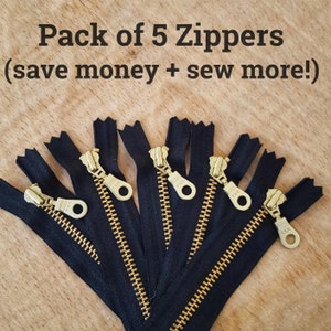 May include: A pack of five black zippers with gold metal pulls. The zippers are arranged in a fan shape on a wooden surface. The text "Pack of 5 Zippers (save money + sew more!)" is visible at the top of the image.