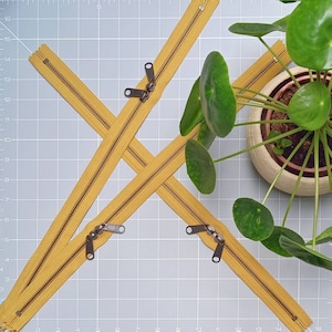May include: Four yellow zipper pulls with metal sliders on a grey grid background. The zipper pulls are arranged in a criss-cross pattern. A potted plant with green leaves is in the background.