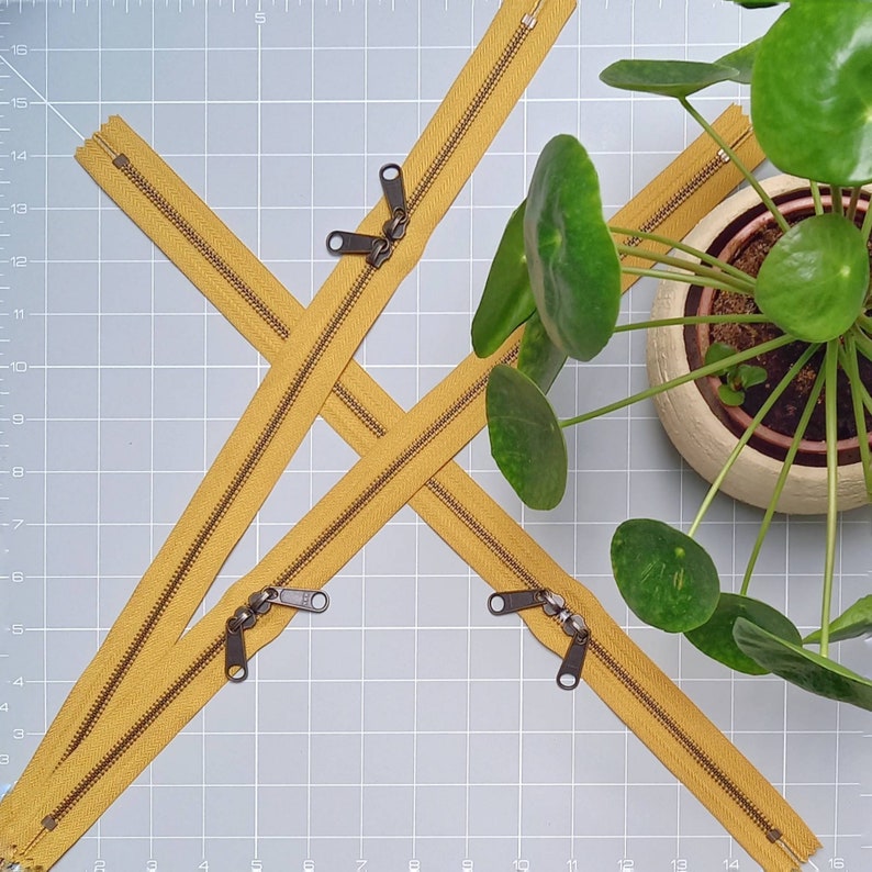 May include: Four yellow zipper pulls with metal sliders on a grey grid background. The zipper pulls are arranged in a criss-cross pattern. A potted plant with green leaves is in the background.