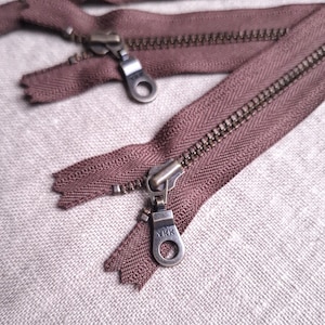 Pode incluir: Close-up de vários zíperes marrons com puxadores de metal. Os zíperes estão abertos, revelando os dentes e a marca YKK no puxador. O tecido é de cor neutra e texturizado.